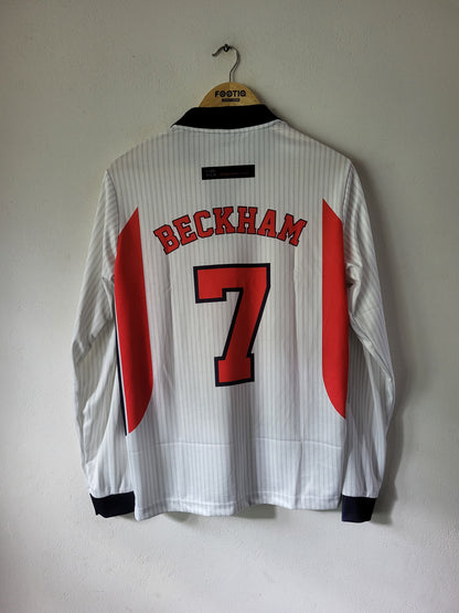 A white long-sleeve football jersey with 'BECKHAM' and the number '7' in red on the back, hanging on a wooden hanger against a plain wall.