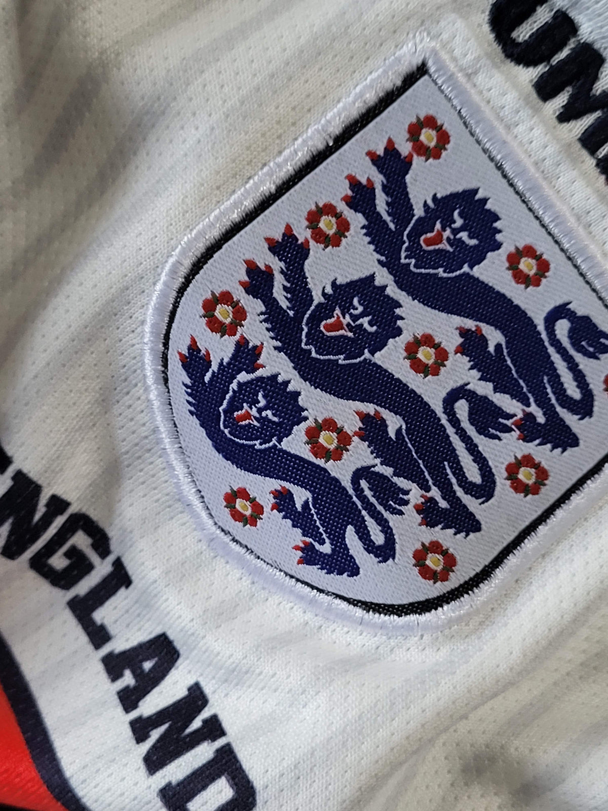 Close-up of a white England soccer jersey showing the embroidered national crest with three lions and red roses, along with partial black lettering.