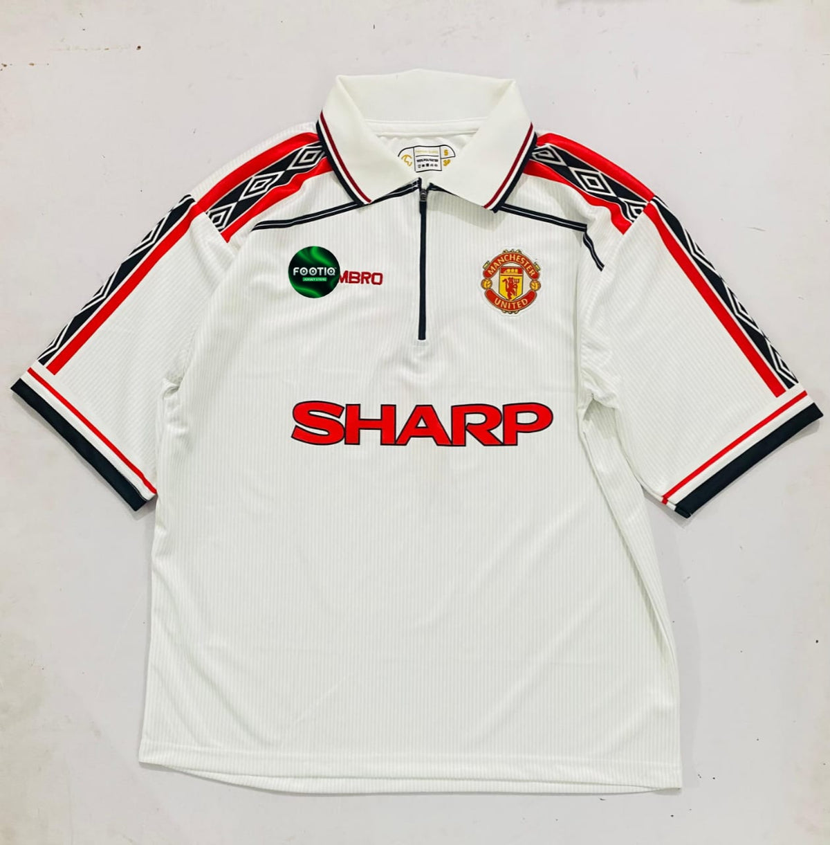 A vintage white Manchester United short-sleeve polo shirt with a half-zip collar, featuring the red 'SHARP' sponsor logo across the chest, the Manchester United crest, and a green 'Fosterr Hero' badge. The sleeves have black and red geometric patterns.
