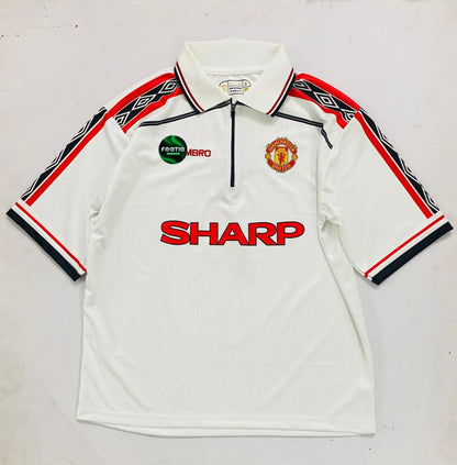 A vintage white Manchester United short-sleeve polo shirt with a half-zip collar, featuring the red 'SHARP' sponsor logo across the chest, the Manchester United crest, and a green 'Fosterr Hero' badge. The sleeves have black and red geometric patterns.