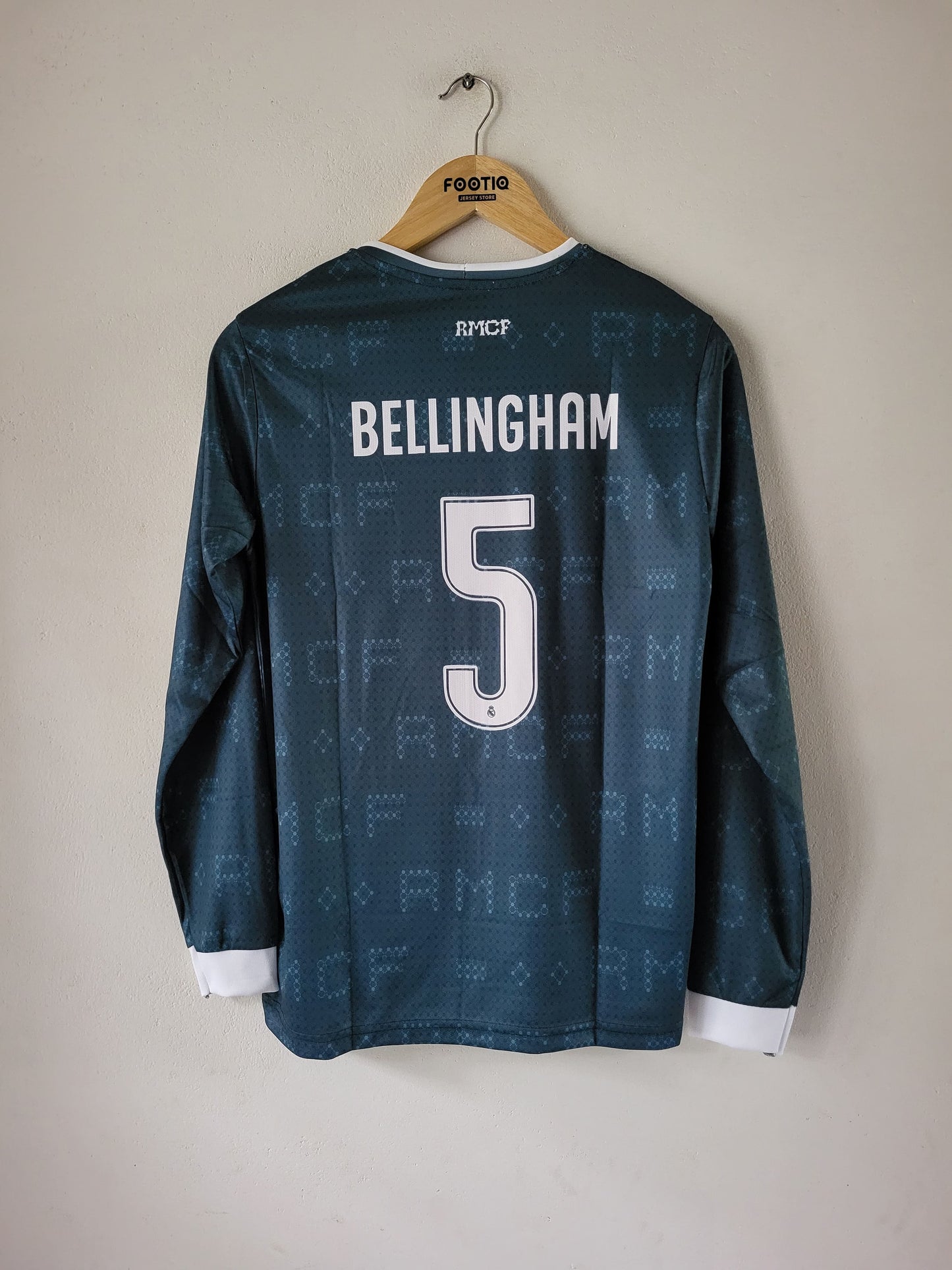 A dark teal long-sleeve soccer jersey with 'BELLINGHAM' and the number '5' printed on the back, hanging on a wooden hanger against a plain beige wall.