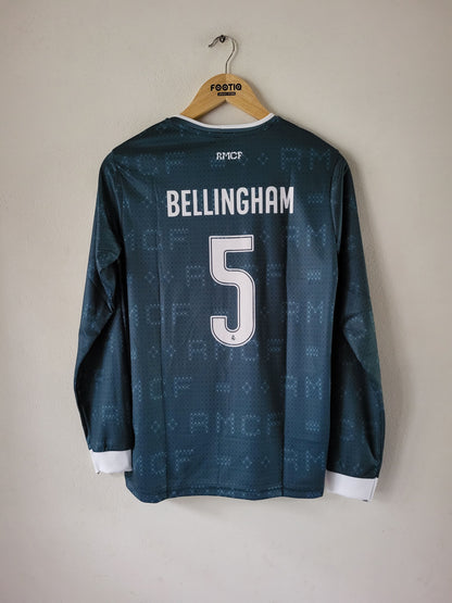 A dark teal long-sleeve soccer jersey with 'BELLINGHAM' and the number '5' printed on the back, hanging on a wooden hanger against a plain beige wall.