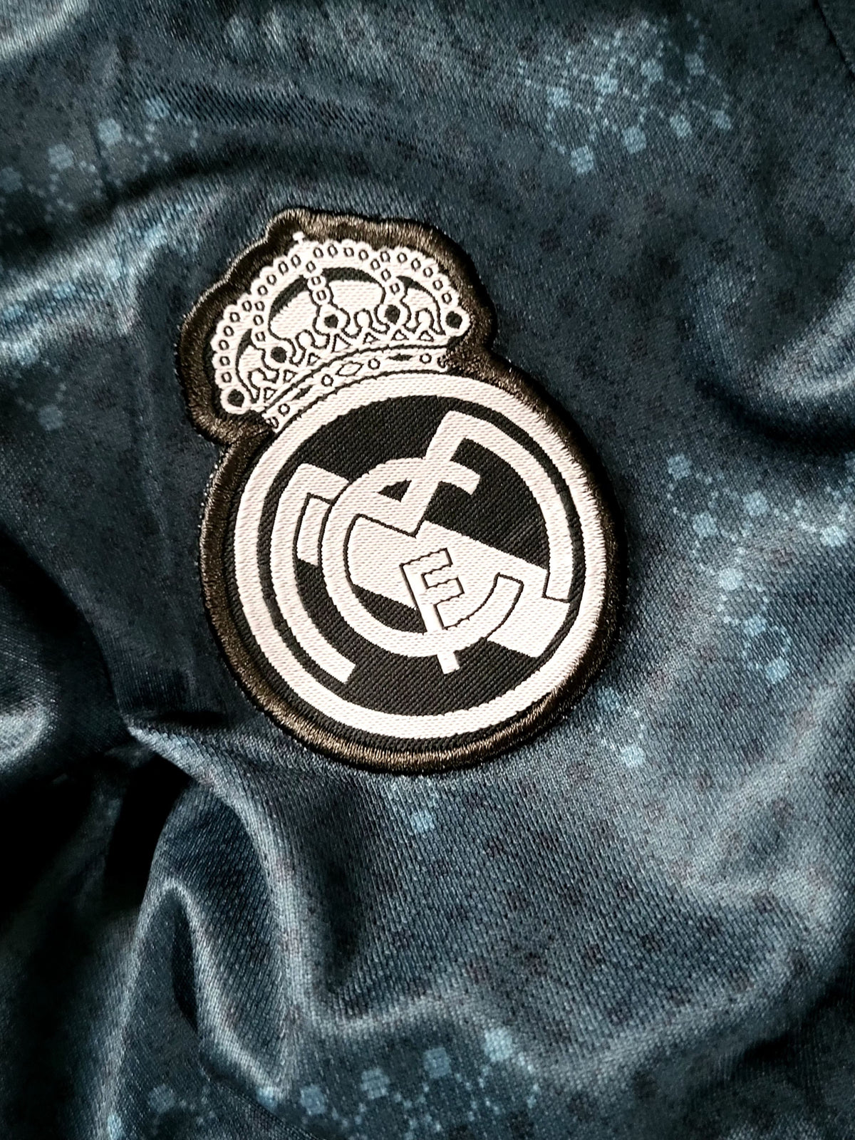 A close-up of a Real Madrid embroidered patch on a dark, textured fabric background.