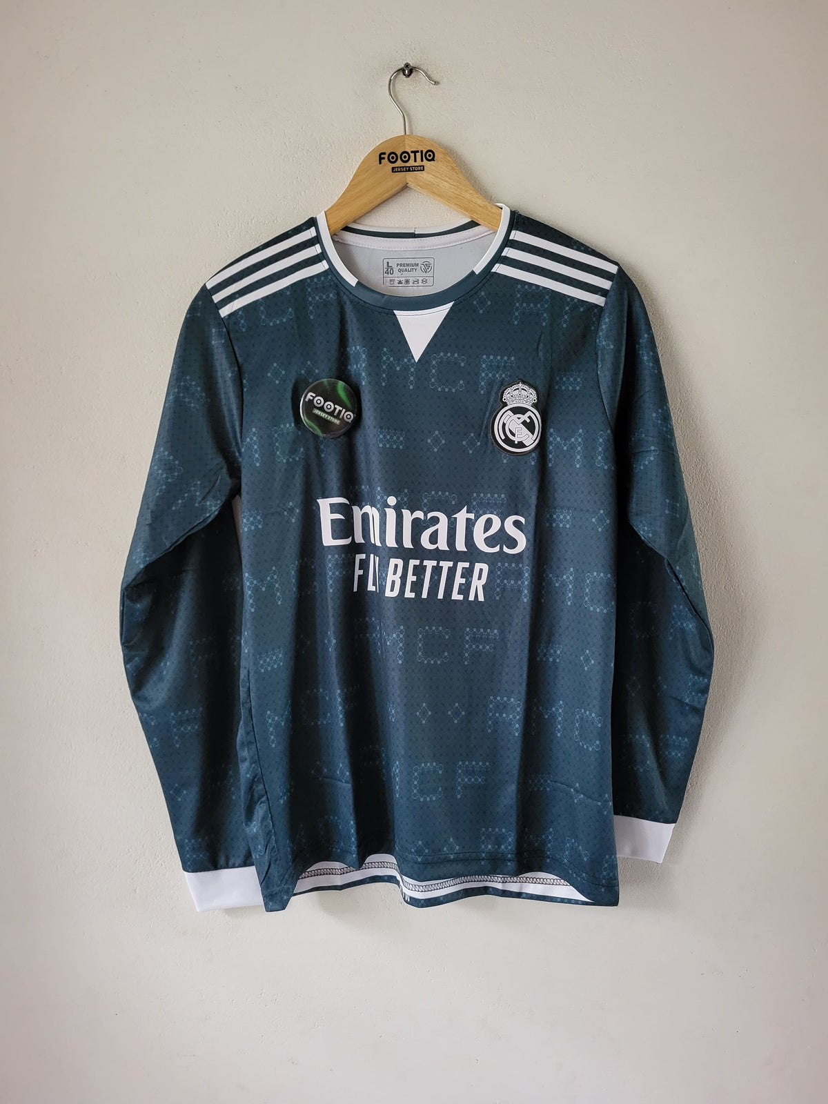 A dark teal Real Madrid soccer jersey with white accents, hanging on a wooden hanger against a plain beige wall. The jersey features the Emirates Fly Better sponsor logo, the Real Madrid crest, and Adidas shoulder stripes.