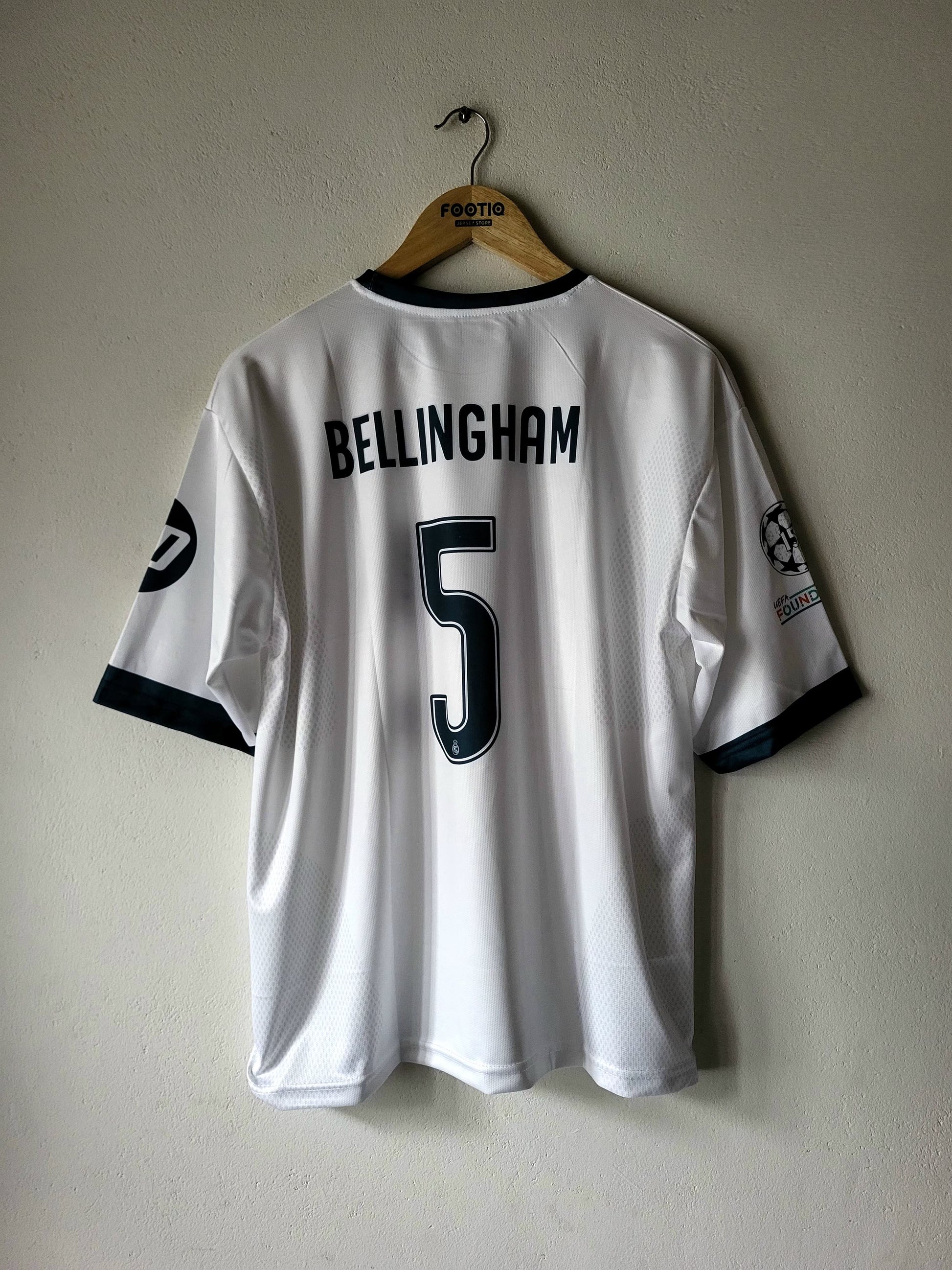 A white Real Madrid soccer jersey with 'BELLINGHAM' and the number '5' on the back, hanging on a wooden hanger against a textured beige wall.