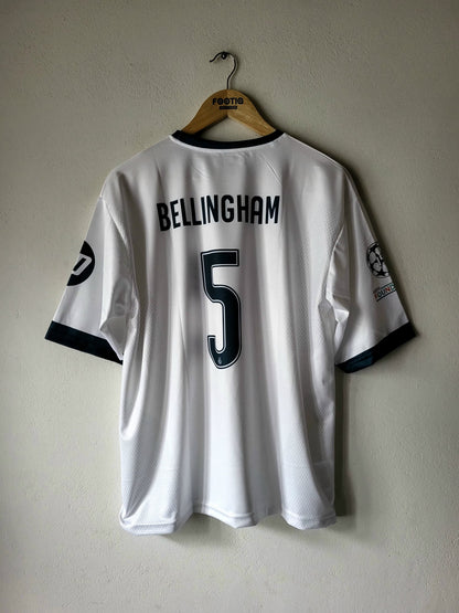 A white Real Madrid soccer jersey with 'BELLINGHAM' and the number '5' on the back, hanging on a wooden hanger against a textured beige wall.