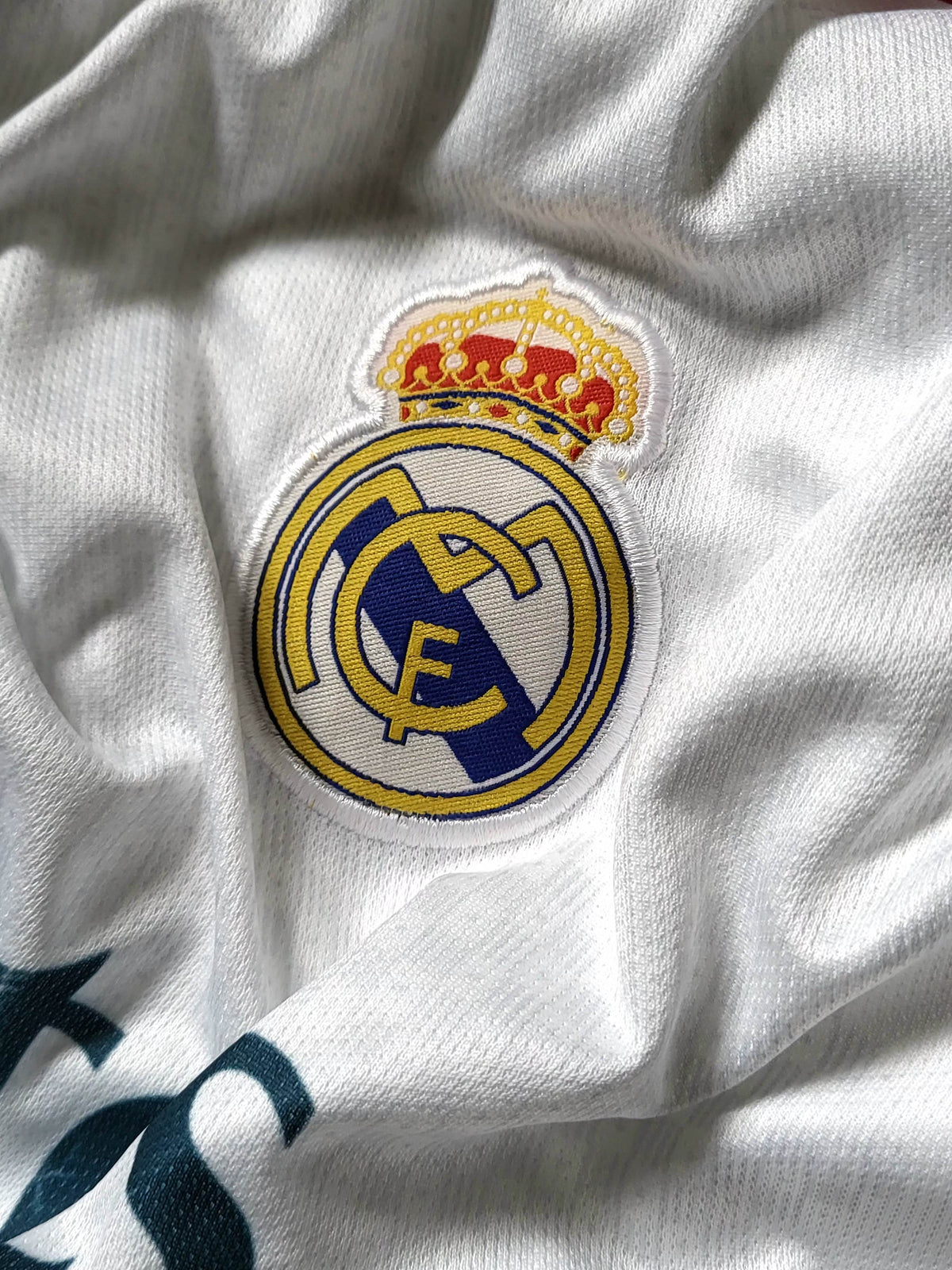 A close-up of a white Real Madrid soccer jersey showing the embroidered club crest with a gold crown on the chest.