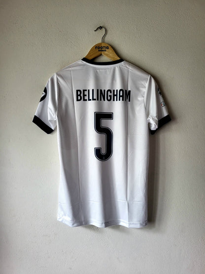 White football jersey with 'BELLINGHAM' and number '5' printed in black on the back, hanging on a wooden hanger against a textured beige wall.