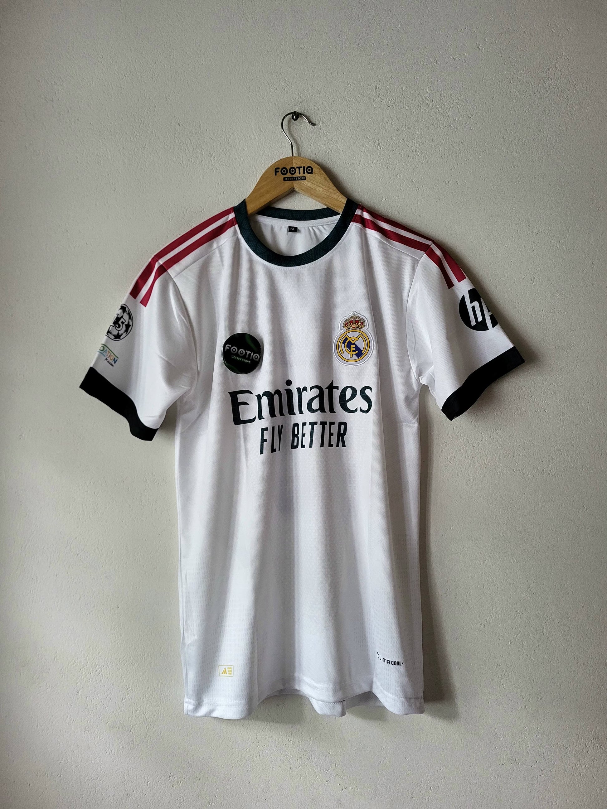 A white Real Madrid soccer jersey with black and red accents, hanging on a wooden hanger against a plain wall. The jersey features the Emirates Fly Better sponsor logo, the Real Madrid crest, and UEFA Champions League patches on the sleeves.