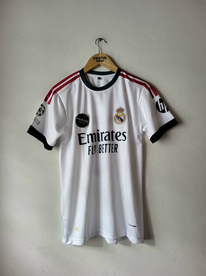 A white Real Madrid soccer jersey with black and red accents, hanging on a wooden hanger against a plain wall. The jersey features the Emirates Fly Better sponsor logo, the Real Madrid crest, and UEFA Champions League patches on the sleeves.