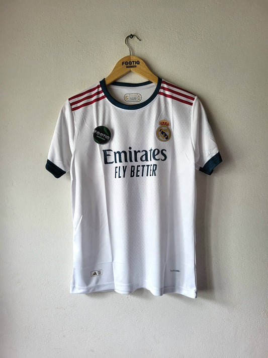 A white Real Madrid soccer jersey with navy blue trim and red shoulder stripes, hanging on a wooden hanger against a plain beige wall. The jersey features the Emirates Fly Better sponsor logo and the Real Madrid crest.