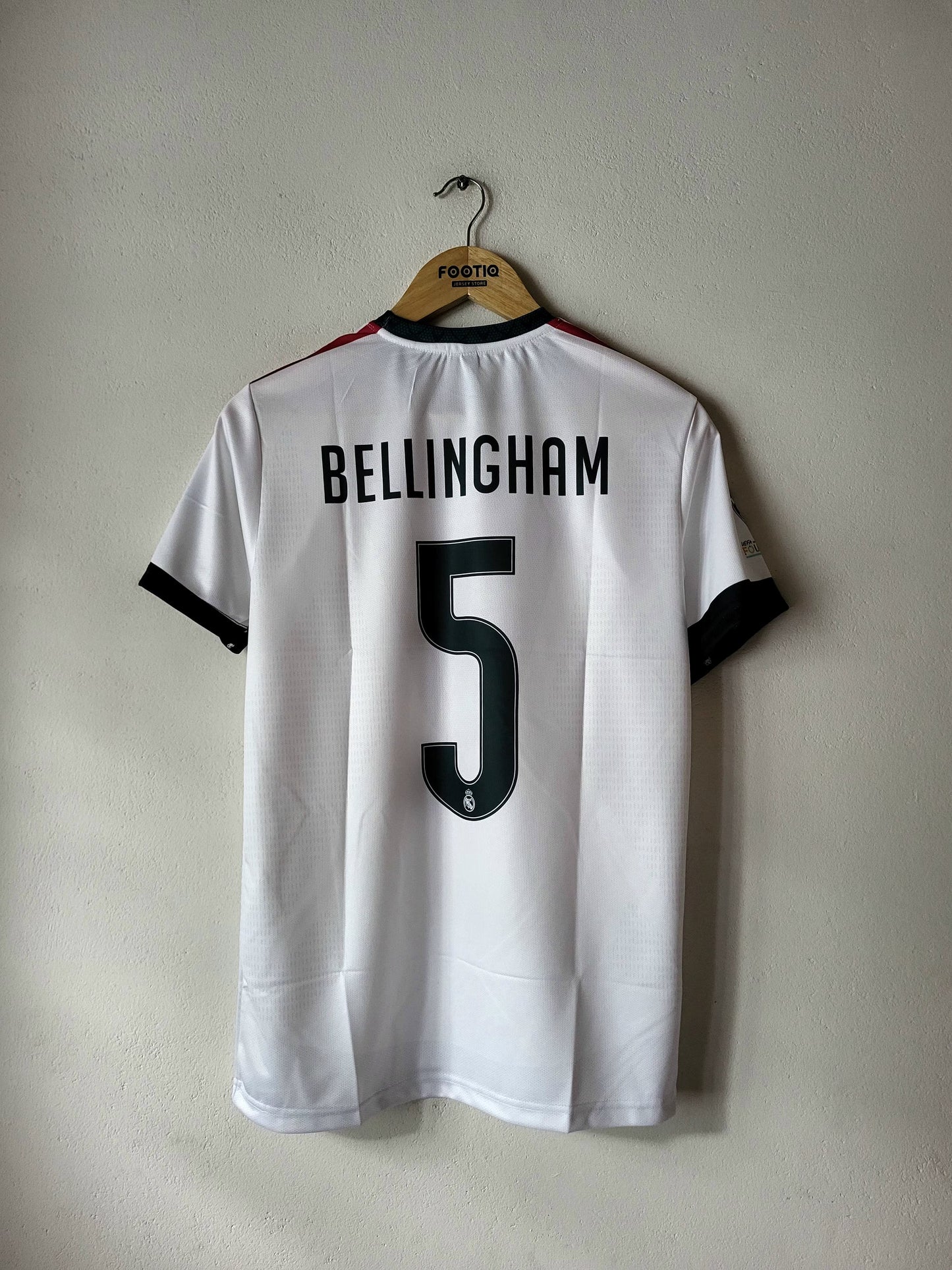 A white short-sleeve soccer jersey hanging on a wooden hanger against a plain light gray wall. The back of the shirt shows the name BELLINGHAM in black block letters above a large black number 5.