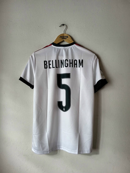 A white short-sleeve soccer jersey hanging on a wooden hanger against a plain light gray wall. The back of the shirt shows the name BELLINGHAM in black block letters above a large black number 5.