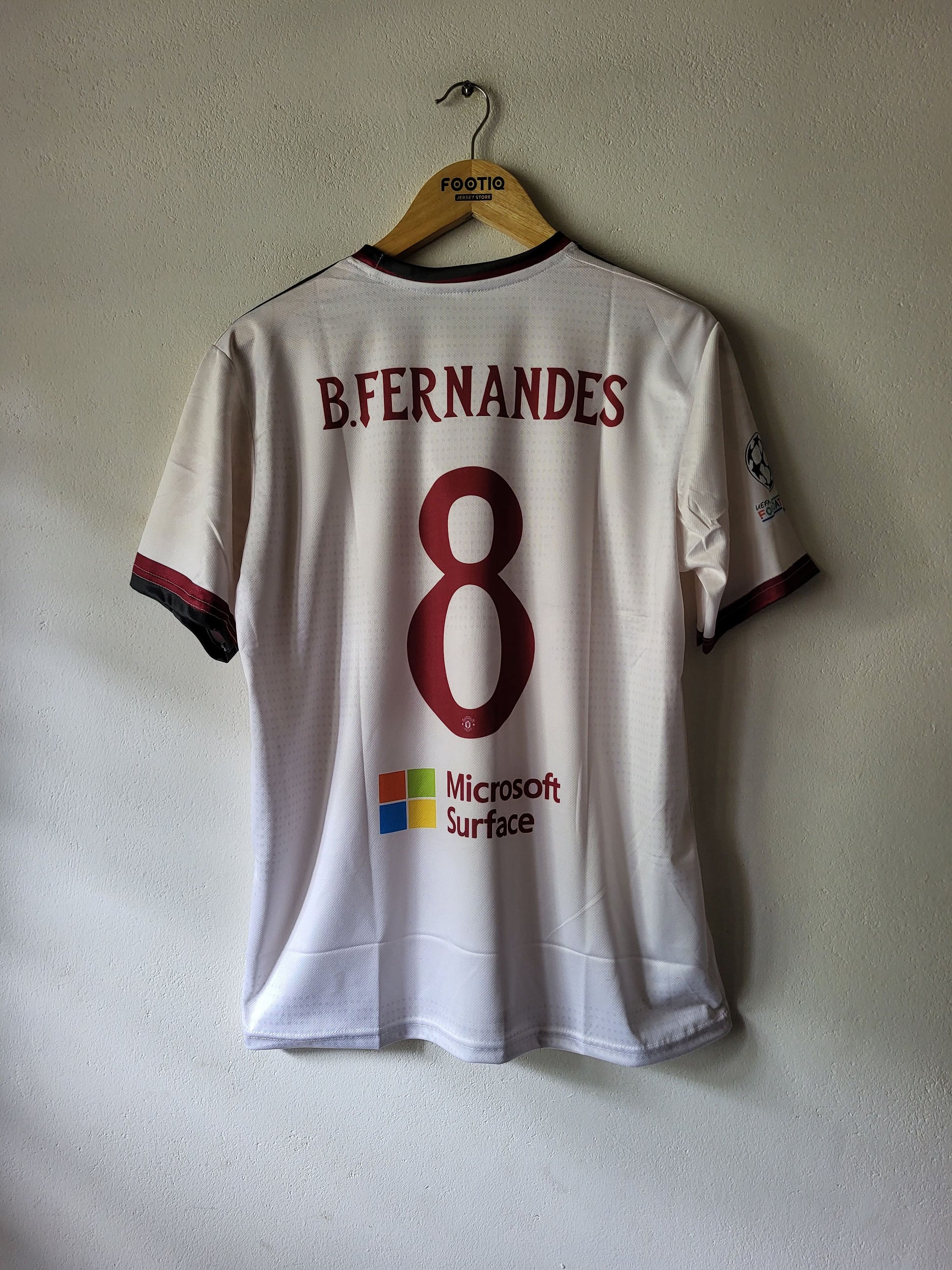 A white Manchester united soccer jersey with the back of the number 8, featuring 'B. FERNANDES' and a Microsoft Surface logo, hanging on a wooden hanger against a textured wall.