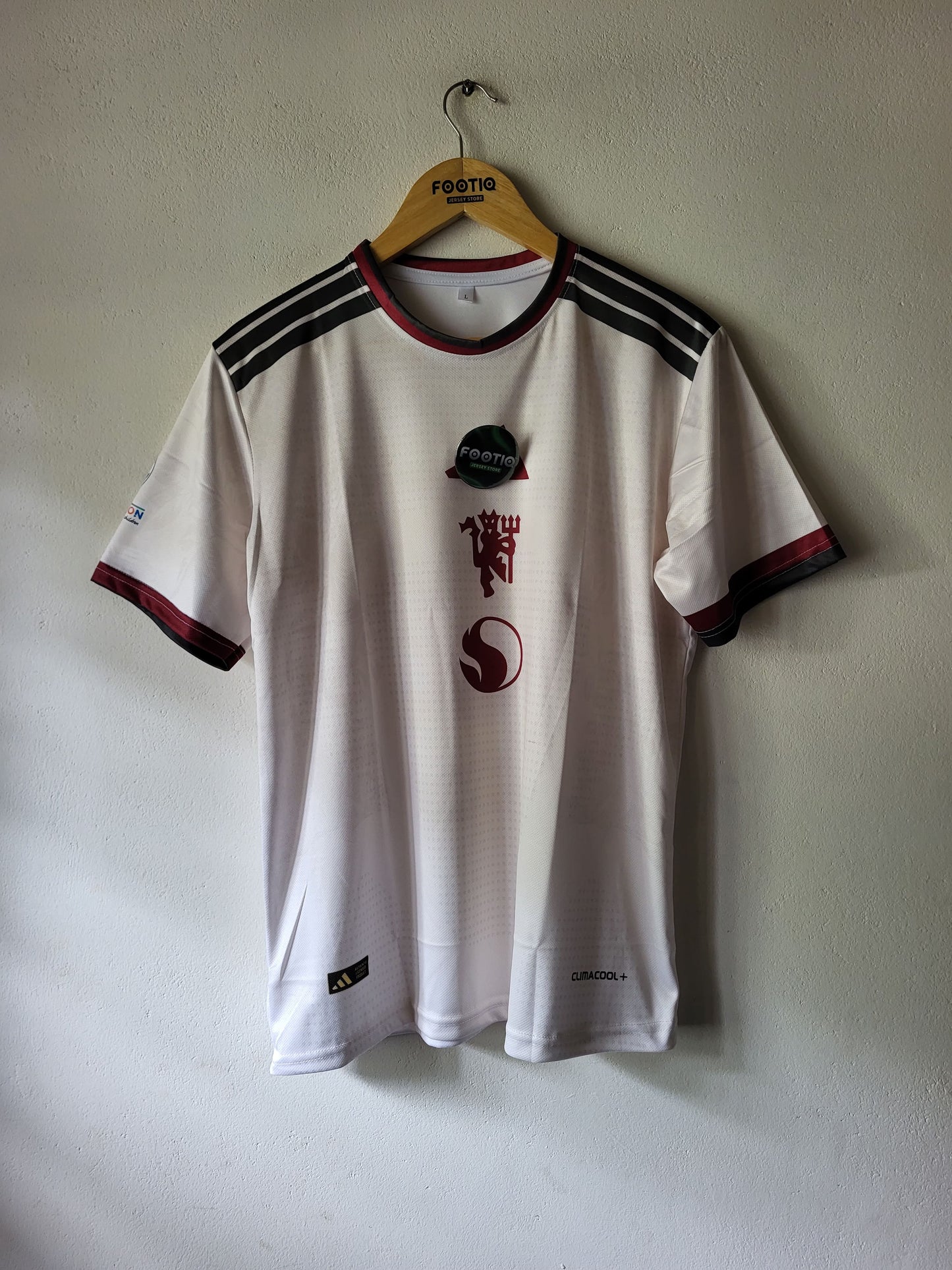 A white soccer jersey with black shoulder stripes and red trim, hanging on a wooden hanger against a plain wall. The jersey features the Manchester united club crest and sponsor logos on the front.