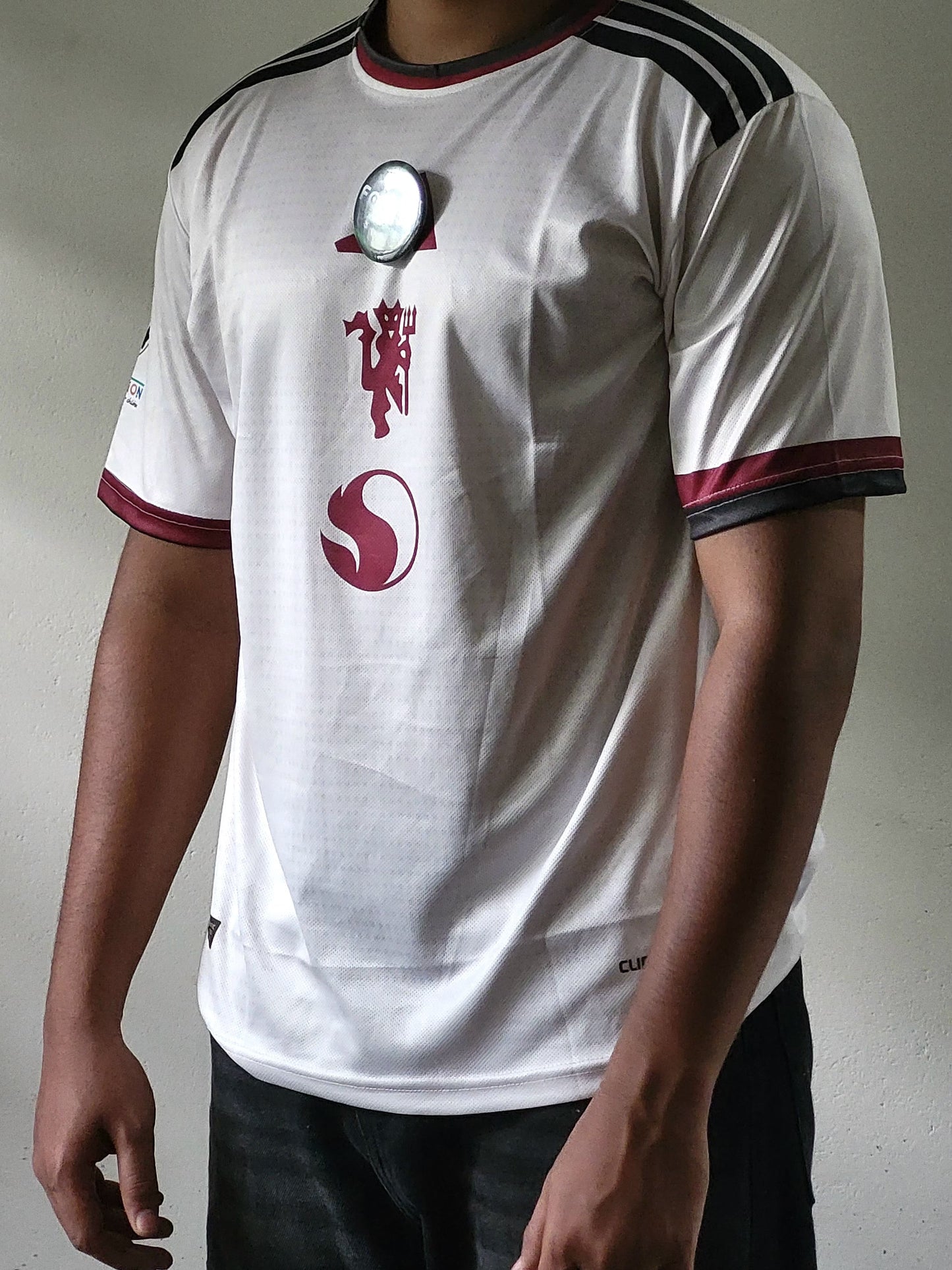 A white short-sleeve soccer jersey with black shoulder stripes, maroon trim on the collar and sleeve cuffs, and a maroon Manchester United crest on the chest, worn by a model.