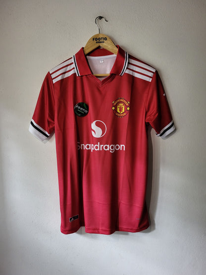 A red Manchester United short-sleeve soccer jersey hanging on a wooden hanger against a plain wall. The jersey features white shoulder stripes, the Manchester United crest on the chest, a black sponsor patch, and a white Snapdragon sponsor logo.