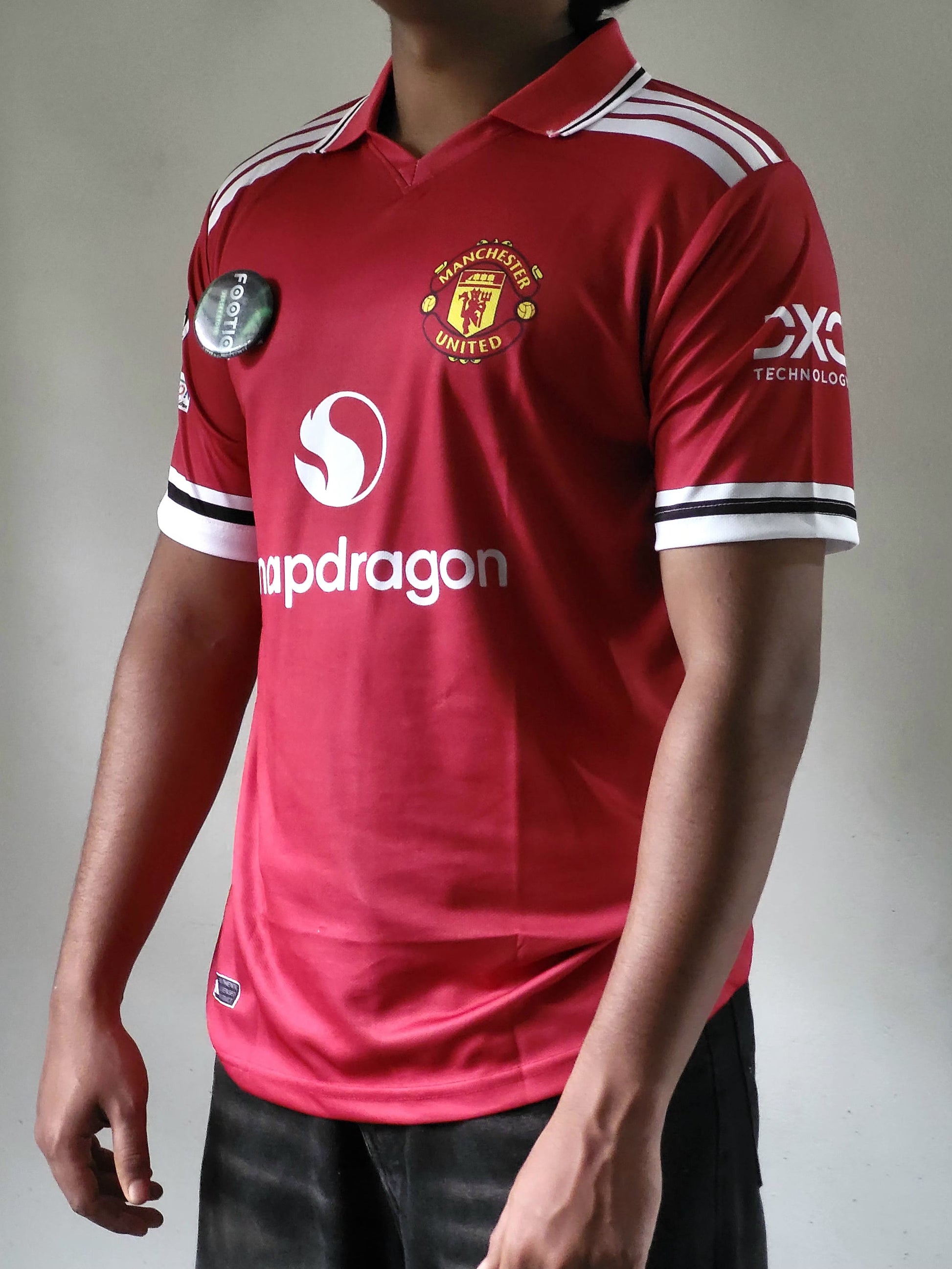 A person wearing a red Manchester United soccer jersey with white and black sleeve stripes, white shoulder stripes, and visible sponsor logos, photographed against a plain light gray background.
