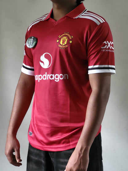 A person wearing a red Manchester United soccer jersey with white and black sleeve stripes, white shoulder stripes, and visible sponsor logos, photographed against a plain light gray background.