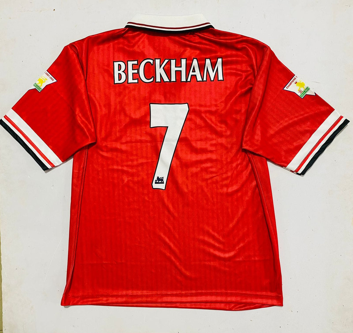 A red Manchester United Beckham jersey with the name 'BECKHAM' and number '7' printed in white on the back, laid flat against a light background.