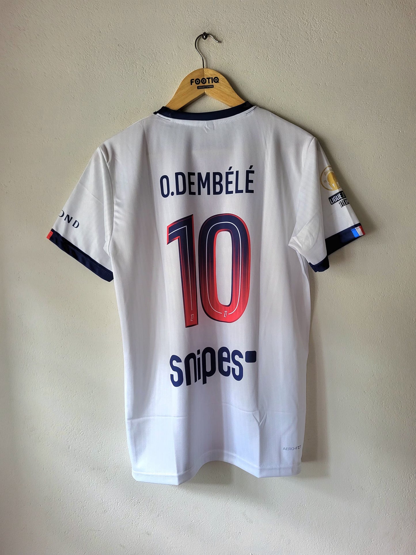 White O. Dembélé number 10 soccer jersey with navy and red accents, hanging on a wooden hanger against a beige wall.
