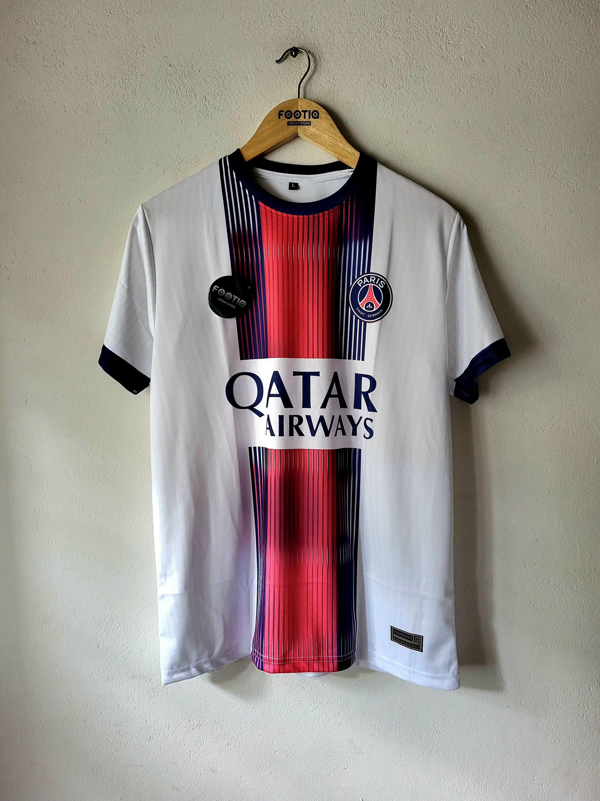 A Paris Saint-Germain home soccer jersey hanging on a wooden hanger against a textured beige wall. The jersey is white with a central red and navy blue vertical stripe pattern, featuring the 'Qatar Airways' sponsor logo on the chest and the club's crest on the right side.