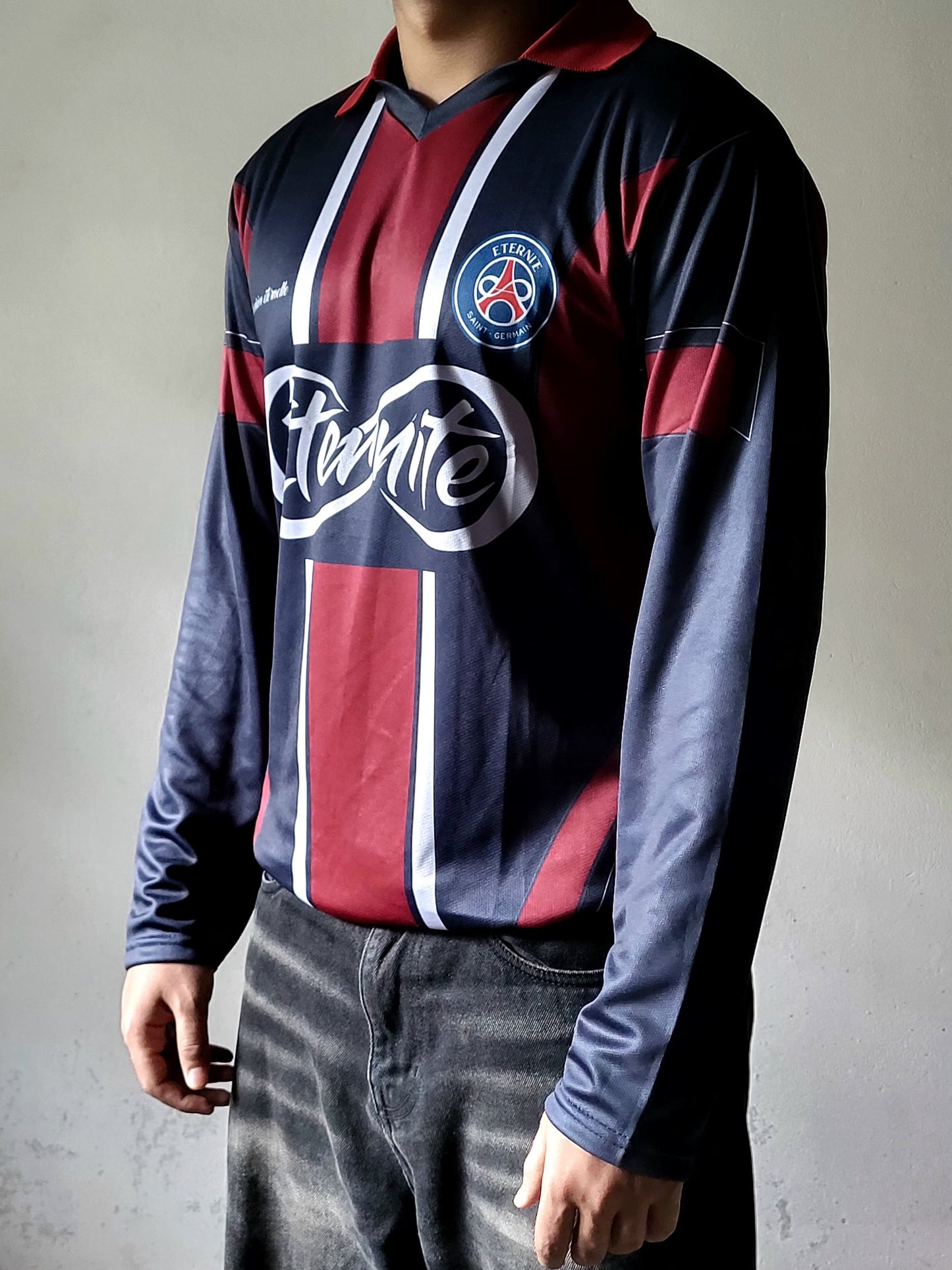 A person wearing a long-sleeve Paris Saint-Germain football jersey with a dark navy base, red vertical stripes, white accents, and a large 'Cristale' sponsor logo across the chest, paired with faded black jeans against a neutral gray background.