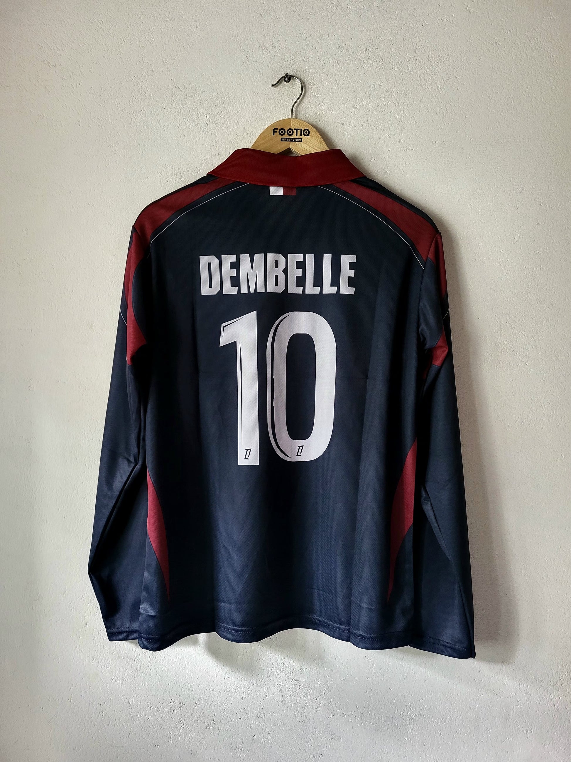 A dark navy blue long-sleeve soccer jersey with 'DEMBELLE' and the number '10' printed in white on the back, hanging on a wooden hanger against a plain off-white wall.