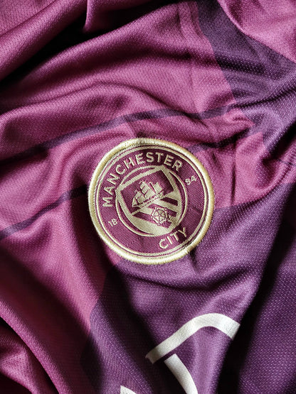 A close-up of a Manchester City soccer jersey in a deep purple color, featuring the embroidered club crest in gold and white on the chest.