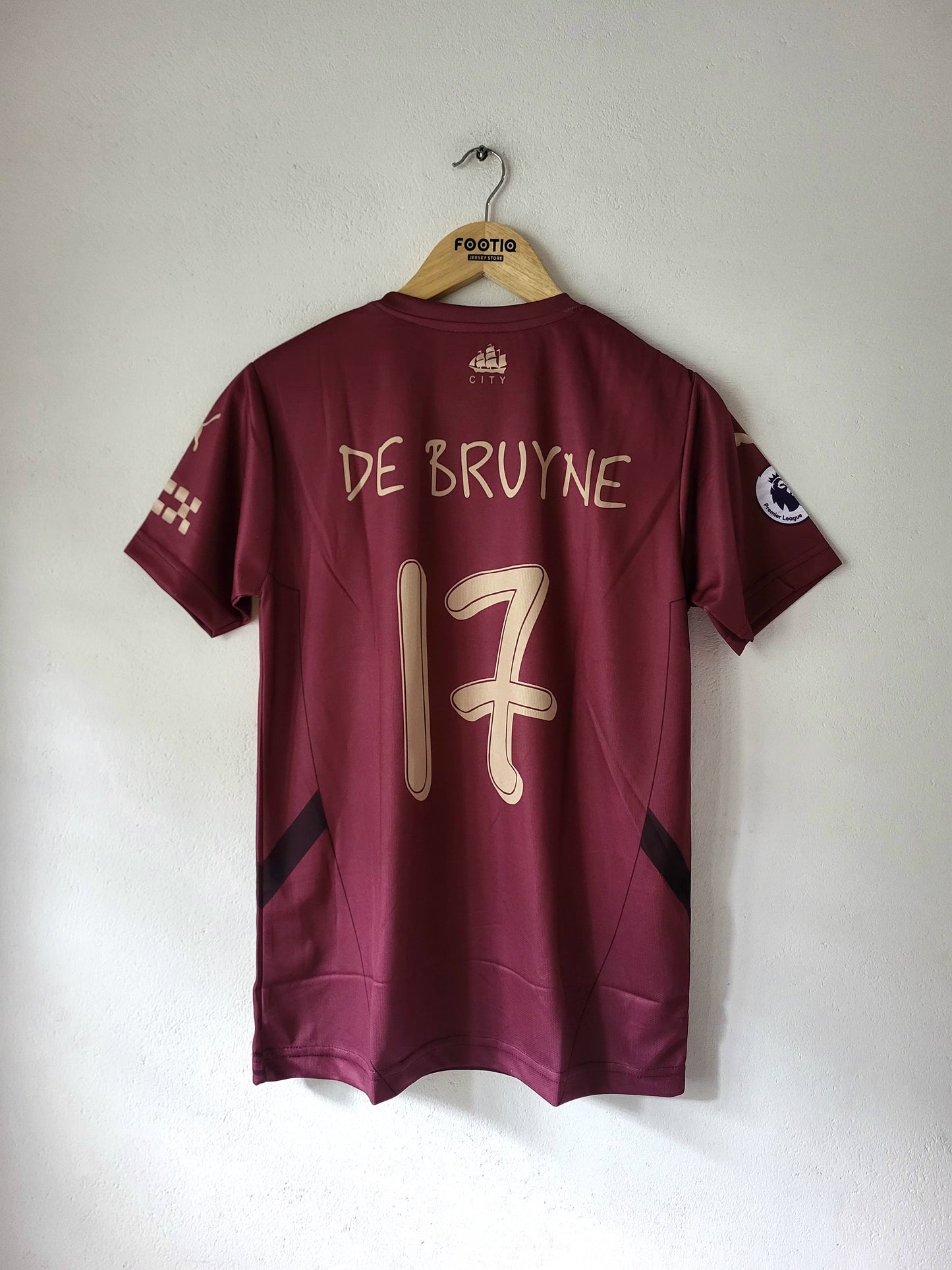 A maroon short-sleeve soccer jersey hanging on a wooden hanger against a plain white wall. The back of the jersey shows 'DE BRUYNE' and the number '17' in light beige/gold lettering, with a Premier League patch on the sleeve.