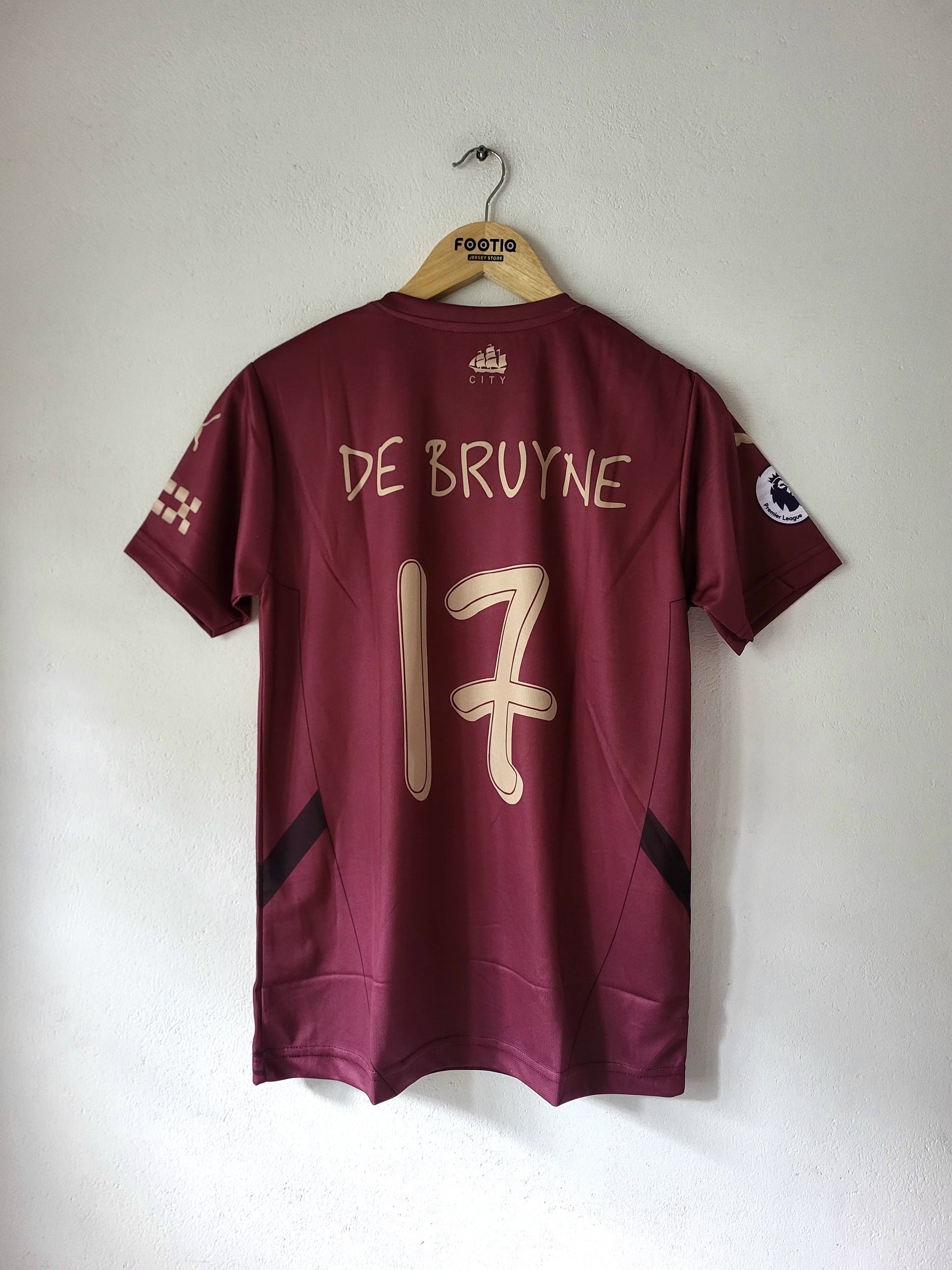 A maroon short-sleeve soccer jersey hanging on a wooden hanger against a plain white wall. The back of the jersey shows 'DE BRUYNE' and the number '17' in light beige/gold lettering, with a Premier League patch on the sleeve.