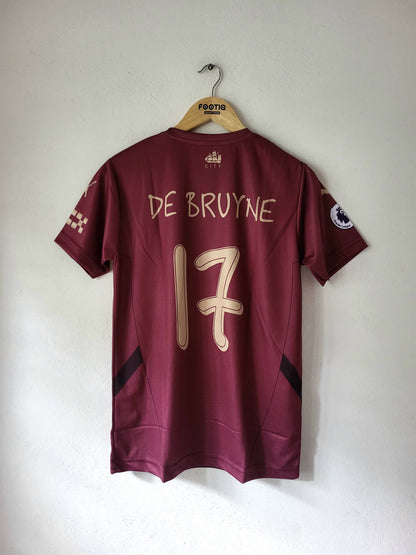 A maroon short-sleeve soccer jersey hanging on a wooden hanger against a plain white wall. The back of the jersey shows 'DE BRUYNE' and the number '17' in light beige/gold lettering, with a Premier League patch on the sleeve.