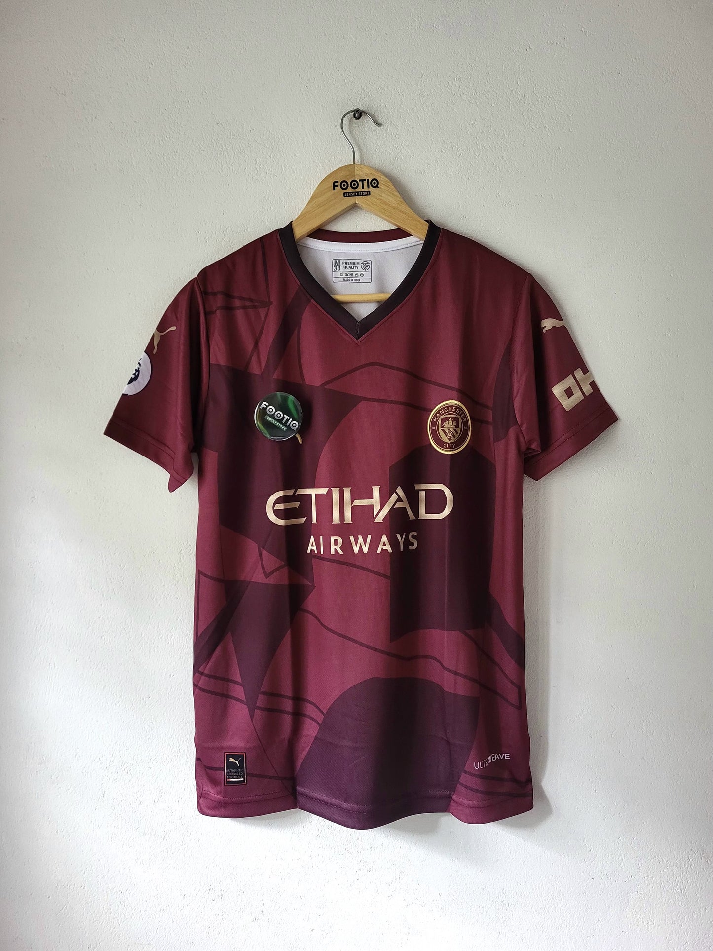 A maroon Manchester City football jersey with gold and white sponsor logos, hanging on a wooden hanger against a plain white wall.