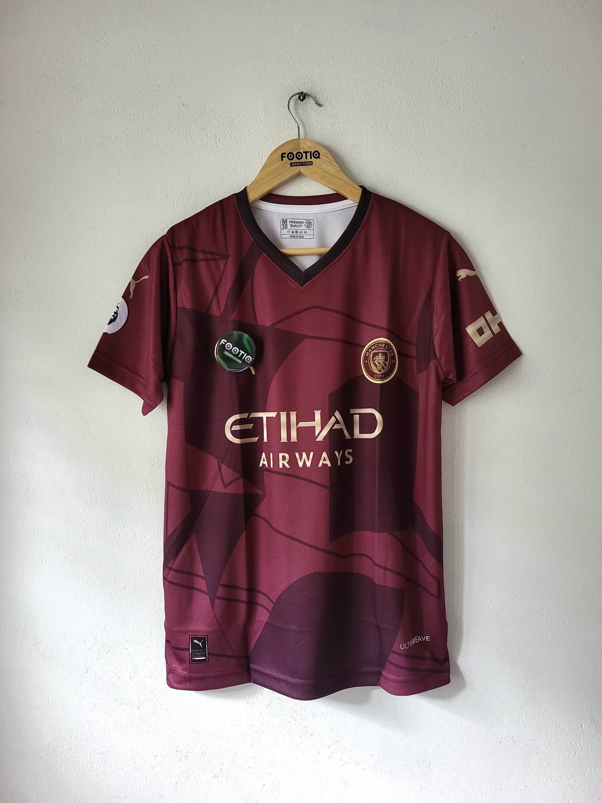 A maroon Manchester City football jersey with gold and white sponsor logos, hanging on a wooden hanger against a plain white wall.