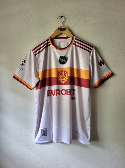 A white AS Roma soccer jersey with red and yellow stripes, hanging on a wooden hanger against a textured beige wall. The jersey features the AS Roma crest, Eurobet Live sponsor logo, and Adidas branding.