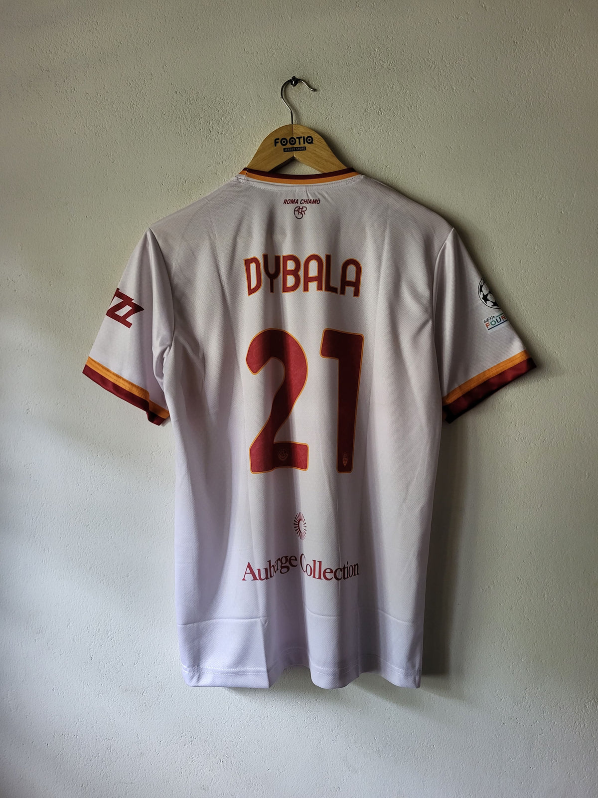 Back view of a white AS Roma football jersey hanging on a wooden hanger against a plain wall. The jersey has 'DYBALA' and the number '21' in red on the back, with AS Roma crest and sponsor logos visible.