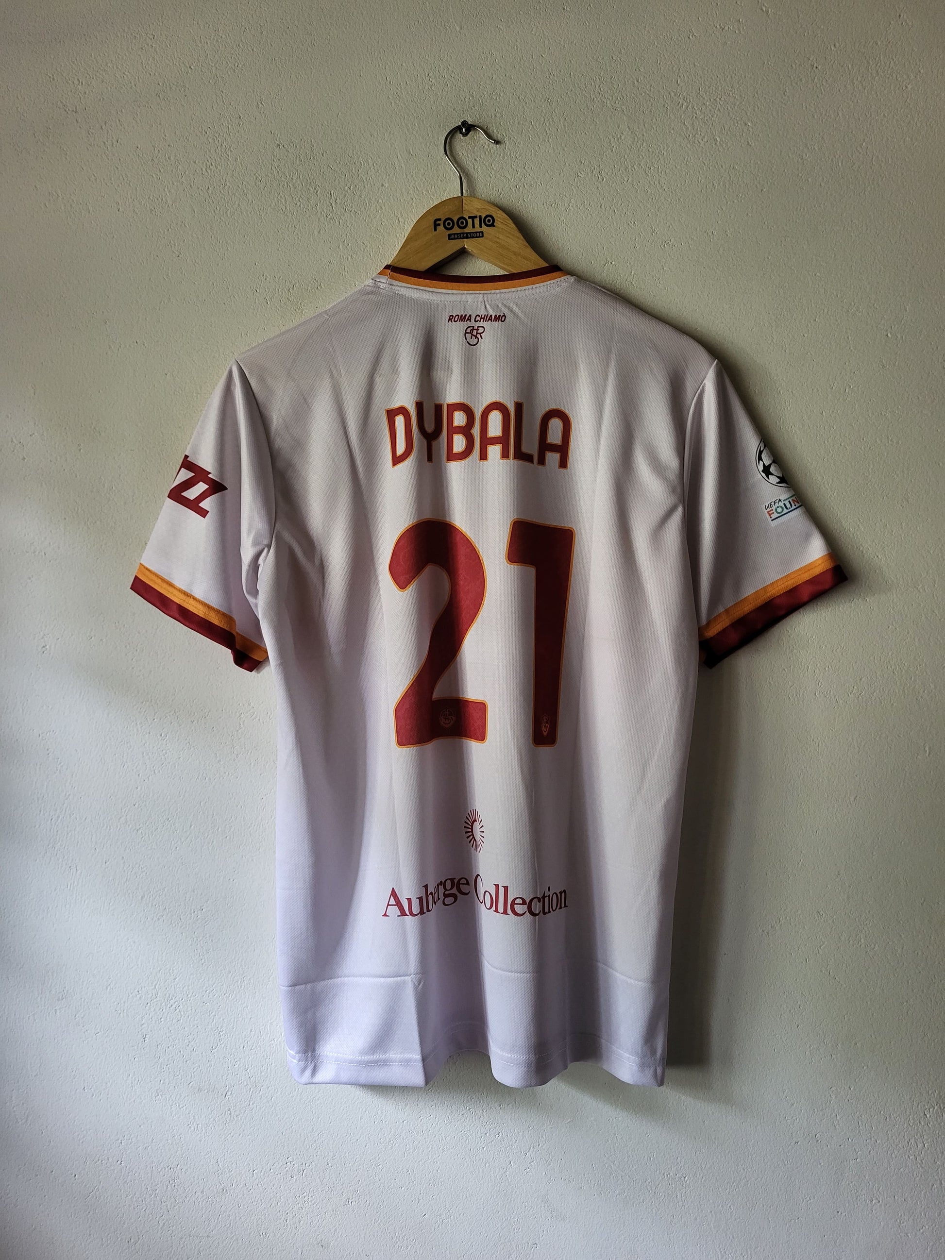 Back view of a white AS Roma football jersey hanging on a wooden hanger against a plain wall. The jersey has 'DYBALA' and the number '21' in red on the back, with AS Roma crest and sponsor logos visible.