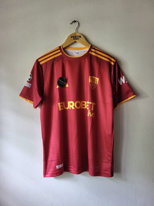 A red AS Roma soccer jersey with yellow accents hangs on a wooden hanger against a light gray wall. The jersey features the yellow Eurobet live sponsor logo on the front, the club crest on the left chest, and yellow shoulder stripes.