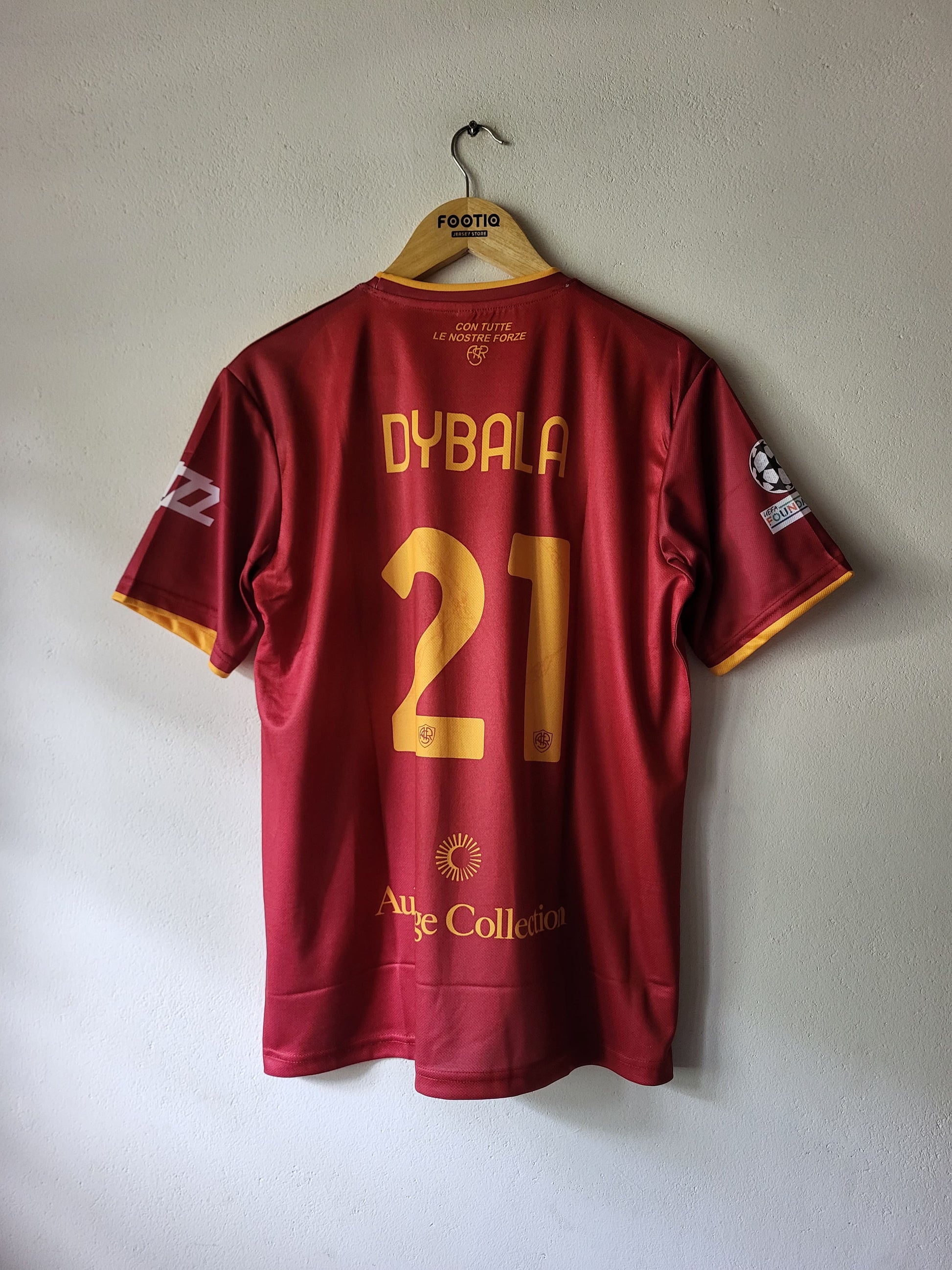 A red and gold AS Roma football jersey with 'DYBALA 21' printed on the back, hanging on a wooden hanger against a light gray wall.