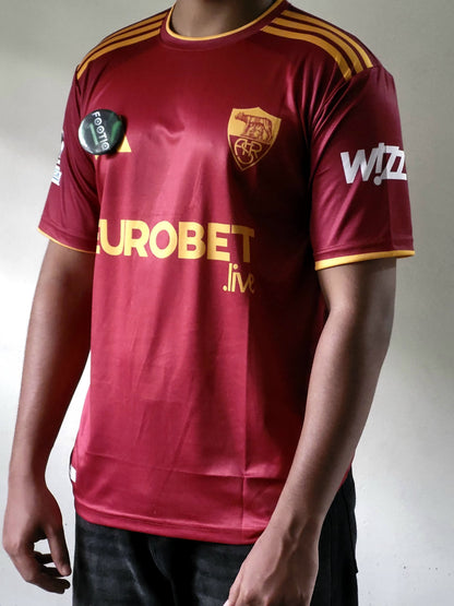 As Roma short-sleeve football jersey with gold shoulder stripes, a gold crest on the chest, and sponsor logos, worn by a model against a plain light background.
