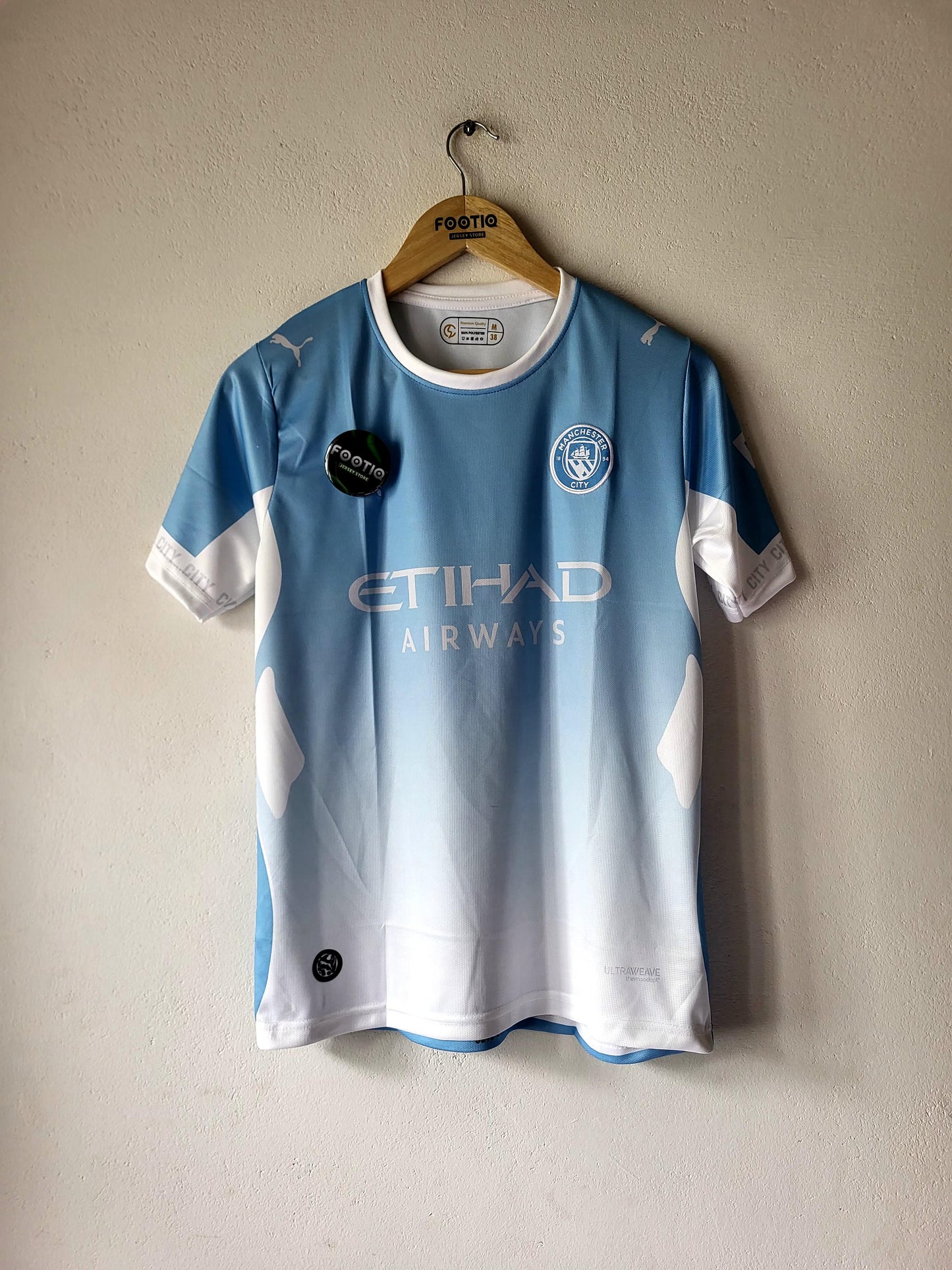A light blue and white gradient Manchester City football jersey hanging on a wooden hanger against a textured beige wall. The jersey features the Etihad Airways sponsor logo on the front, the Manchester City crest on the left chest, and Puma branding.