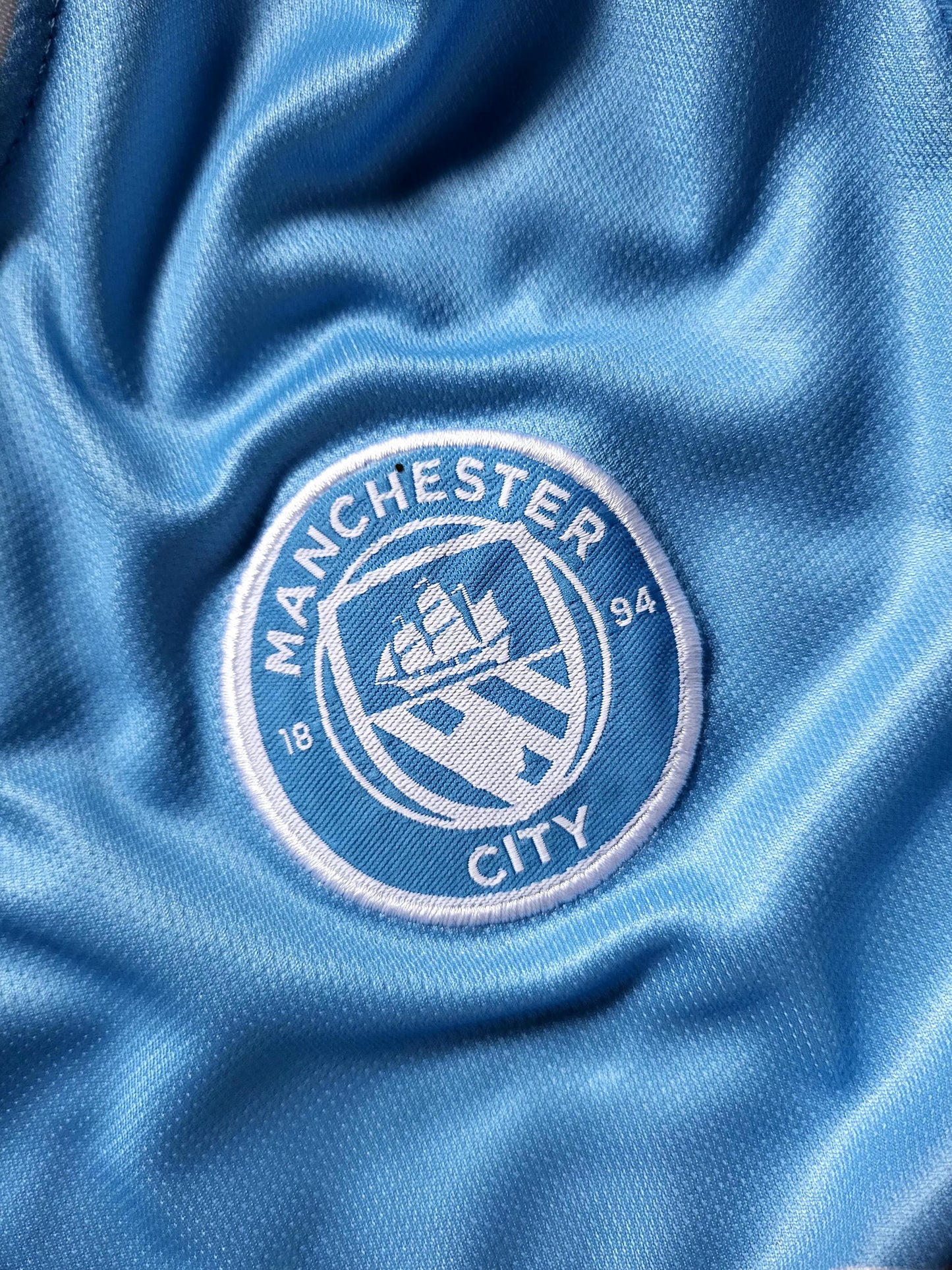 Close-up of a light blue Manchester City soccer jersey fabric with the embroidered club crest in white and blue.
