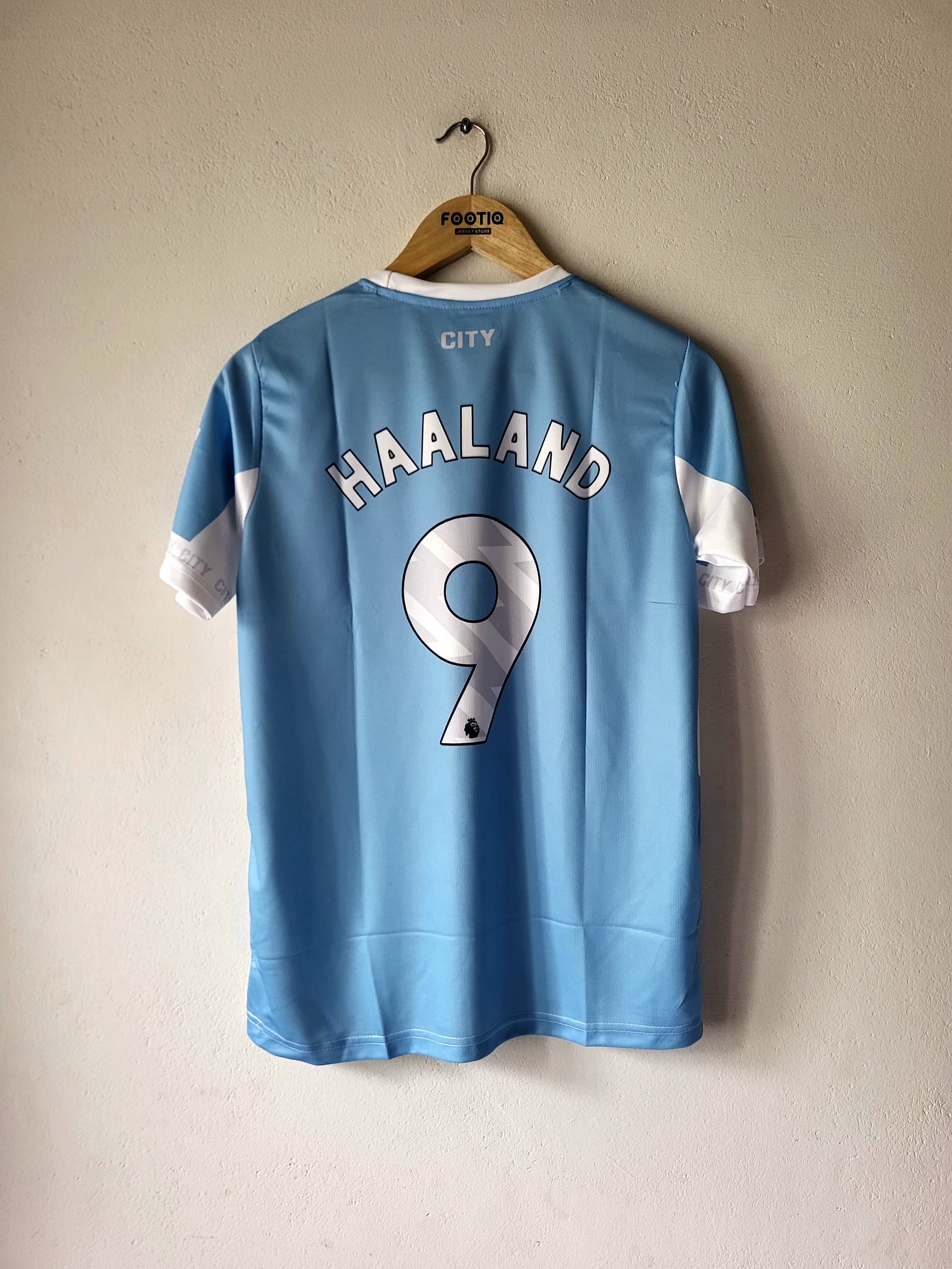 A light blue Manchester City football jersey with 'HAALAND' and the number '9' printed on the back, hanging on a wooden hanger against a textured beige wall.