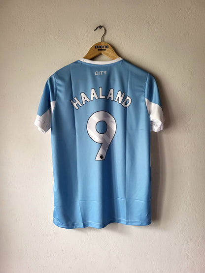 A light blue Manchester City football jersey with 'HAALAND' and the number '9' printed on the back, hanging on a wooden hanger against a textured beige wall.