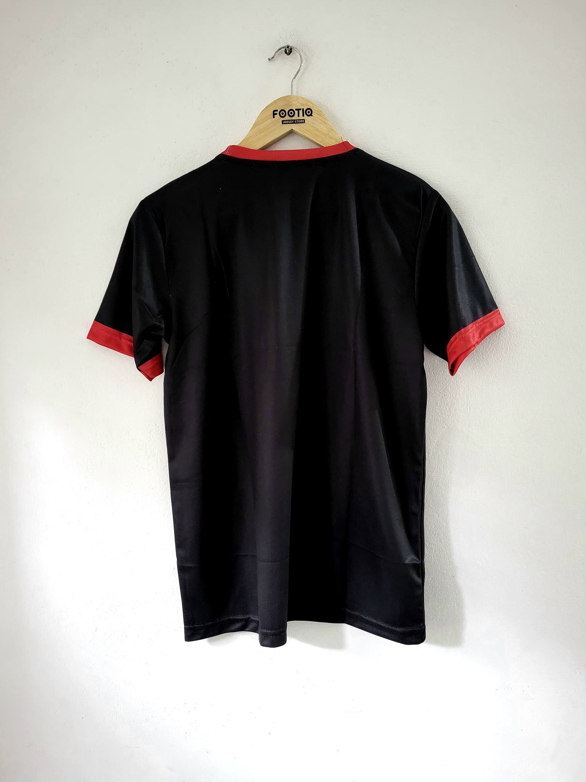 A black short-sleeve athletic jersey with red trim on the collar and sleeve cuffs, hanging on a wooden hanger against a plain white wall.