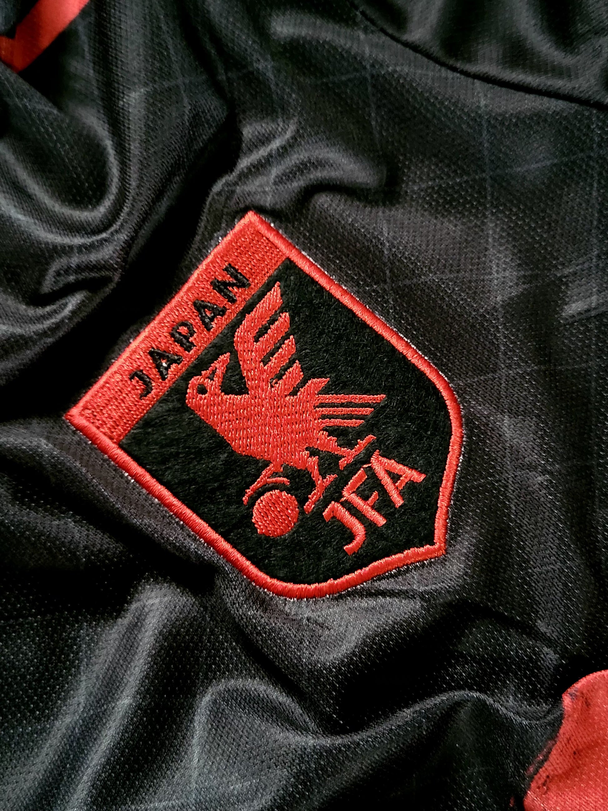 Close-up of a black soccer jersey with an embroidered red and black Japan JFA crest patch on the chest.
