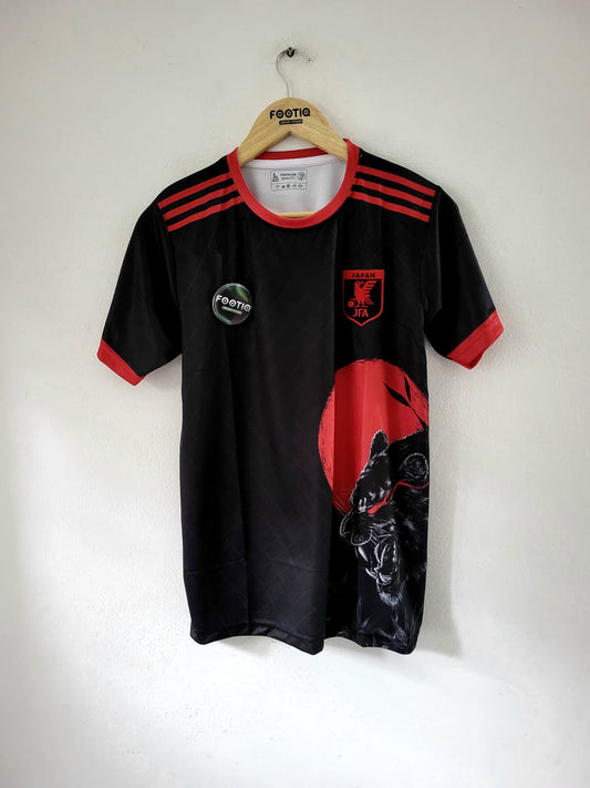 A black short-sleeve soccer jersey with red accents, hanging on a wooden hanger against a plain white wall. The jersey features three red stripes on the shoulders, a red collar, a circular green and white sponsor patch on the right chest, a red crest on the left chest, and a large red graphic with a black animal head on the lower right side.