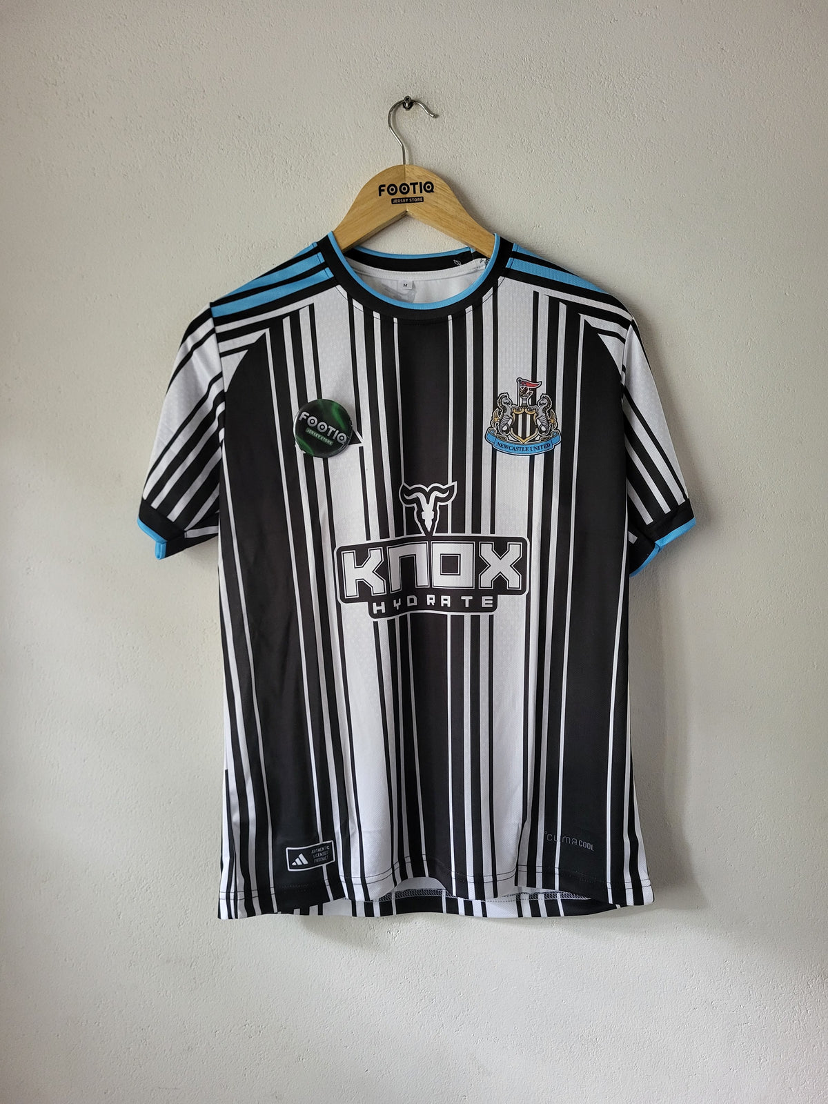 A black and white striped Newcastle United soccer jersey with light blue accents, hanging on a wooden hanger against a plain off-white wall. The jersey features the 'KNOX HYDROTE' sponsor logo on the front.