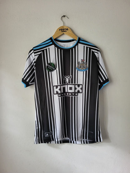 A black and white striped Newcastle United soccer jersey with light blue accents, hanging on a wooden hanger against a plain off-white wall. The jersey features the 'KNOX HYDROTE' sponsor logo on the front.