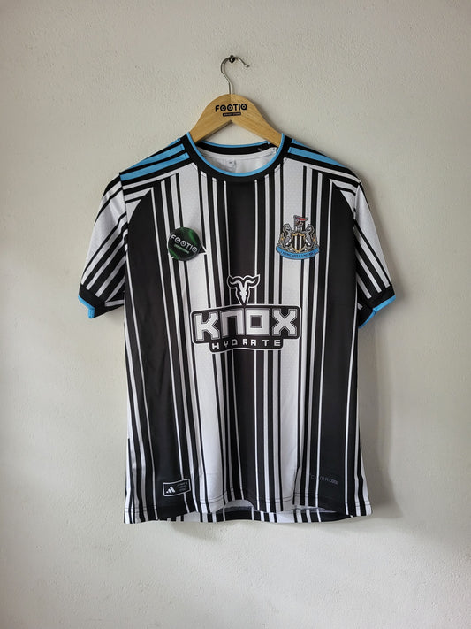 A black and white striped Newcastle United soccer jersey with light blue accents, hanging on a wooden hanger against a plain off-white wall. The jersey features the 'KNOX HYDROTE' sponsor logo on the front.